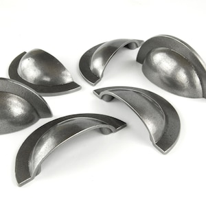 May include: Six antique-style, cast iron drawer pulls in a dark grey colour. The pulls have a textured finish and a curved, cup-like design with a rounded handle. Suitable for cabinets and drawers, these handles measure approximately 10 cm in length.