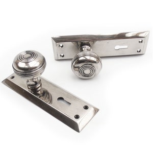 May include: A pair of silver door handles with a keyhole and a round knob with a decorative pattern. The handles are mounted on rectangular plates.