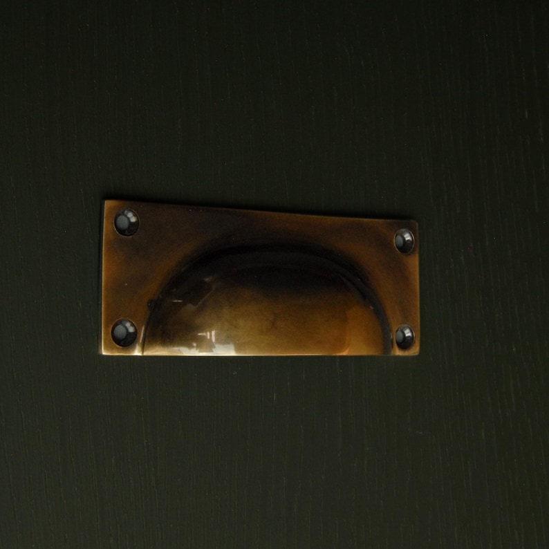 Op de afbeelding: Een rechthoekige antiek messing ladeknop met een gebogen handvat. De knop is gemonteerd op een donkergroen oppervlak en vastgezet met vier kleine schroeven. Het messing heeft een warme, verouderde patina.