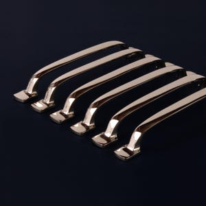 May include: Six polished, gold-toned cabinet handles arranged diagonally on a dark blue background. Each handle has a rectangular base and a curved, bar-shaped grip. The handles are reflective, showing highlights and shadows.