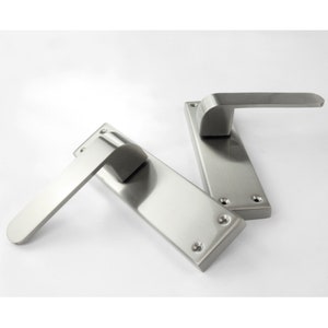 May include: Two brushed steel door handles. Each handle features a rectangular backplate with rounded edges and a curved lever. The handles are made of a shiny, metallic material and are set against a white background.