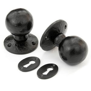May include: Two black door knobs with round handles and oval keyhole covers. The knobs are made of metal and have a hammered finish.