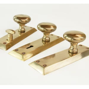 May include: Three brass door handle sets. Each set features a rectangular backplate and a knob or lever. The knobs are oval-shaped. The sets are made of a shiny, golden-coloured metal and are arranged against a white background.