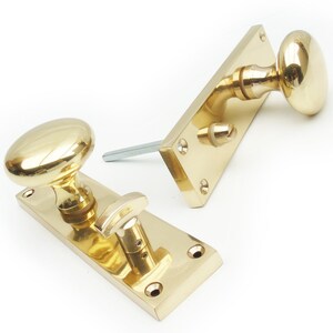 May include: Two polished brass door knobs with oval-shaped handles and a square backplate. The knobs are mounted on a flat surface.