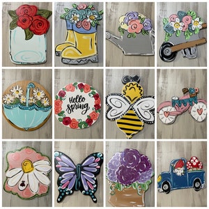 May include: A collection of colorful wooden spring-themed decorations. Designs include a mason jar with red roses, a yellow rain boot with flowers, a watering can, a wheelbarrow, an umbrella, a circle with "Hello Spring", a bee, a bicycle, a daisy, a butterfly, a flower pot, and a truck.