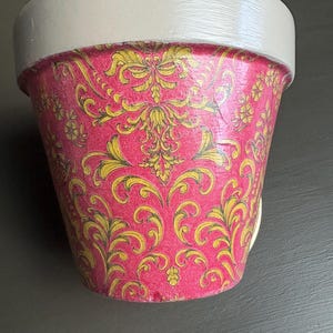 May include: A decorative plant pot featuring a pink base adorned with a gold floral design. The rim is painted white. This pot is designed for plants, adding a decorative element to any room. The pot's height is approximately 15 cm.