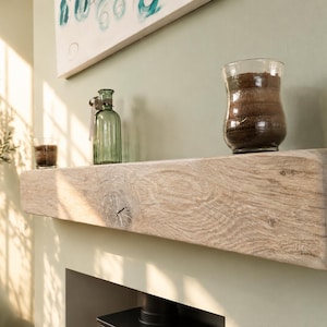 May include: A light-coloured, rustic wooden shelf with a natural grain pattern. The shelf holds a clear glass vase filled with dark material, a small glass, and a green glass bottle. The shelf is mounted on a light green wall.