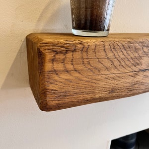 May include: A rustic wooden shelf made from reclaimed oak, with a natural wood finish. The shelf is mounted on a white wall and has a glass with a dark liquid inside sitting on top.