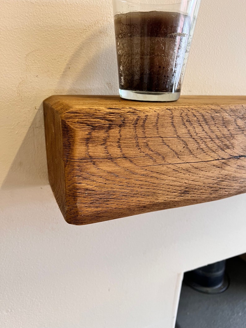 May include: A rustic wooden shelf made from reclaimed oak, with a natural wood finish. The shelf is mounted on a white wall and has a glass with a dark liquid inside sitting on top.