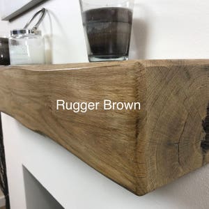 May include: A rustic, Rugger Brown wooden mantelpiece. The mantel is a light brown color with visible wood grain and texture. The text "Rugger Brown" is displayed on the front. Decorative candles and a glass jar are on top.