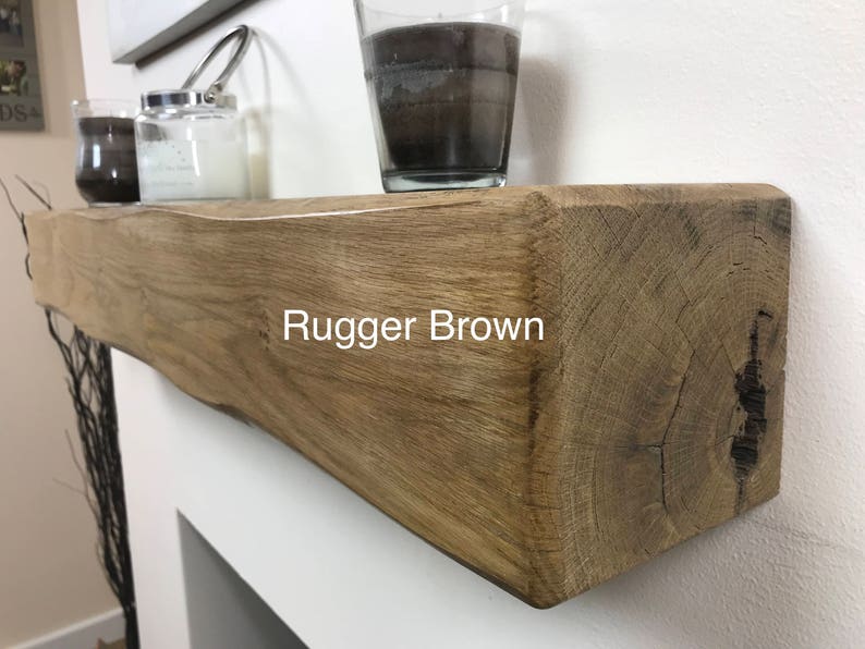 May include: A rustic, Rugger Brown wooden mantelpiece. The mantel is a light brown color with visible wood grain and texture. The text "Rugger Brown" is displayed on the front. Decorative candles and a glass jar are on top.