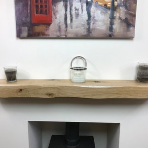 May include: A light-colored, live-edge wooden shelf with a natural finish. The shelf is mounted above a fireplace and holds three glass candle holders. A painting hangs above the shelf.