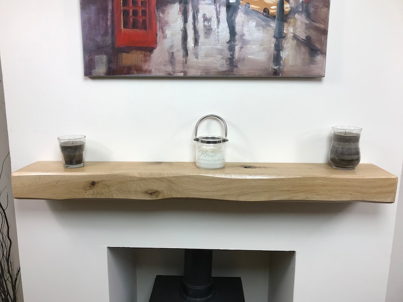 May include: A light-colored, live-edge wooden shelf with a natural finish. The shelf is mounted above a fireplace and holds three glass candle holders. A painting hangs above the shelf.