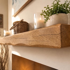 May include: A rustic, solid wood floating shelf with a natural finish. The shelf is mounted on a white wall and holds a small potted plant, a candle in a glass holder, and a woven basket. The shelf's edge has a wavy design.
