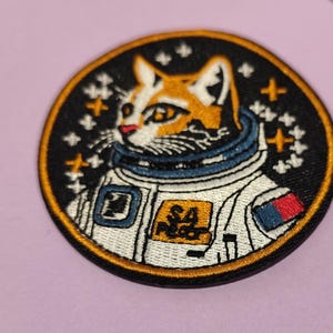 Peut inclure: Écusson brodé circulaire représentant un chat astronaute. Le patch a un fond noir avec une bordure dorée. Le chat est orange et blanc, portant une combinaison spatiale bleue et blanche avec le texte "SA Meow" sur le devant.
