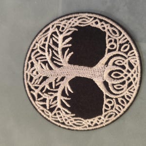 Peut inclure: Un écusson brodé circulaire présentant un motif d'arbre de vie blanc avec des branches et des racines complexes en forme de nœuds sur un fond noir uni. Les bords de l'écusson sont finis par une couture de bordure sombre et serrée.