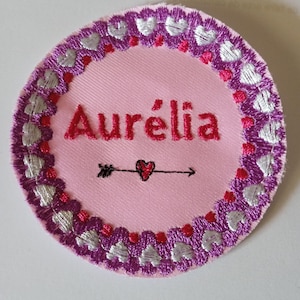 Peut inclure: Un écusson brodé rose circulaire avec le nom "Aurélia" en script rouge. Le patch présente une bordure de cœurs violets et blancs, avec une flèche noire et un motif de cœur sous le nom. Il s'agit probablement d'un article personnalisé.