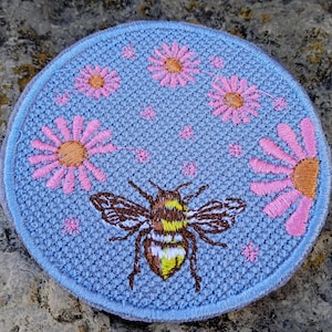 Peut inclure: Un écusson rond brodé avec un fond bleu clair. Il présente une abeille détaillée avec des marques marron et jaune, entourée de marguerites roses et de petites fleurs roses. L'écusson a une bordure bleu clair.