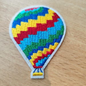 Peut inclure: Écusson brodé en forme de montgolfière. Le ballon est orné de sections rouges, jaunes, bleues et vertes. Il possède une bordure blanche et un petit détail de panier en bas.