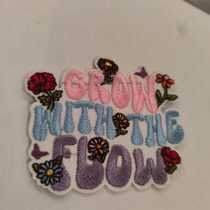 Peut inclure: Un écusson brodé avec le texte "GROW WITH THE FLOW" en fil rose, bleu clair et violet. Le motif est décoré de petites fleurs colorées en rouge, orange et bleu, ainsi que de minuscules papillons violets sur un fond blanc.