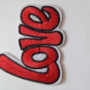 Peut inclure: Écusson brodé avec le mot "love" en rouge, souligné de noir, sur fond blanc. L'écusson présente une police cursive, avec les lettres reliées. L'écusson est probablement destiné aux vêtements ou aux accessoires.
