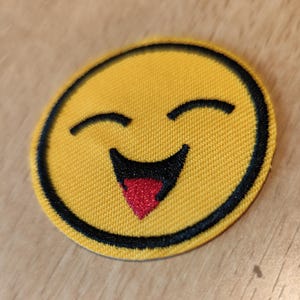 Peut inclure: Un écusson jaune et circulaire représentant un emoji riant. L'emoji a des yeux noirs et un contour noir, avec une langue rouge qui dépasse. L'écusson est en tissu.