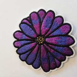 Peut inclure: Patch brodé en forme de fleur, de couleur violette et bleue, avec un centre jaune. La fleur est bordée de noir.