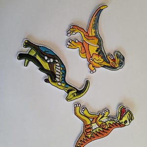 Peut inclure: Trois écussons de dinosaures brodés en jaune, orange et bleu. Les écussons présentent des coutures détaillées et représentent diverses poses de dinosaures, dont une avec la gueule ouverte. Les écussons sont sur fond blanc.