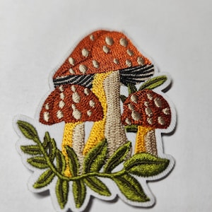 Peut inclure: Un écusson brodé représentant trois champignons aux chapeaux orange à pois blancs, aux pieds jaunes et beiges et aux lamelles noires. Les champignons sont entourés de feuilles de fougère vertes, le tout bordé de blanc sur fond blanc.