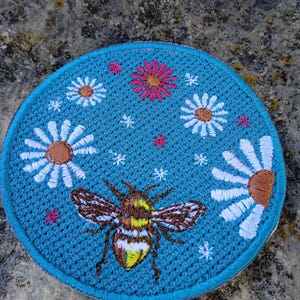 Peut inclure: Un écusson brodé rond avec un fond bleu turquoise. Le patch représente une abeille détaillée avec des marques marron et jaune, entourée de marguerites blanches avec des centres bruns et de petites fleurs roses. Le patch mesure environ 7.6 cm de diamètre.