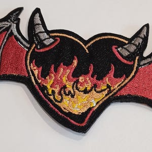 Peut inclure: Un cœur noir avec des flammes rouges et jaunes, entouré d'ailes rouges et grises avec des cornes noires. Le cœur a un contour noir et les ailes ont une bordure noire.