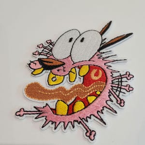 Peut inclure: Écusson brodé représentant Courage le chien peureux avec une expression surprise. Le patch a un contour rose, des yeux blancs et une langue marron. La bouche est rouge avec des dents jaunes et les oreilles sont marron.