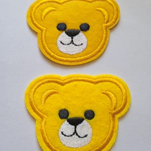 Peut inclure: Deux écussons de nounours jaunes avec des détails brodés. Chaque écusson représente un visage d'ours souriant avec des yeux noirs et un nez noir. Les écussons sont soulignés de coutures jaunes et ont une texture semblable à du feutre.