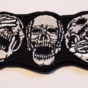 Peut inclure: Trois crânes brodés sur un patch noir. Le crâne du milieu a la bouche ouverte et les crânes de chaque côté se couvrent les yeux et les oreilles avec leurs mains.