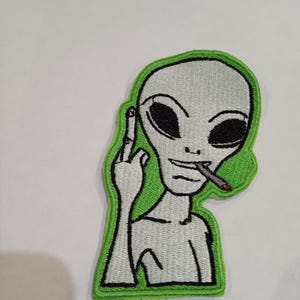 Peut inclure: Patch brodé vert d'un extraterrestre gris faisant un doigt d'honneur et fumant une cigarette.