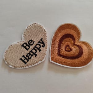 Peut inclure: Deux écussons brodés en forme de cœur. L'un est beige avec les mots "Be Happy" en noir. L'autre est marron avec un motif en spirale. Les deux ont une bordure blanche et sont sur fond blanc.