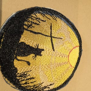 Peut inclure: Patch brodé avec une silhouette de chat noir, une croix et un soleil jaune se levant au-dessus d'une forêt sombre. Le patch est circulaire et a une bordure bleue.