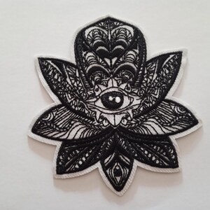 Peut inclure: Écusson brodé noir et blanc en forme de fleur de lotus. Le patch présente des détails complexes, dont un œil au centre et des motifs ornés sur les pétales. Le patch a une bordure blanche.