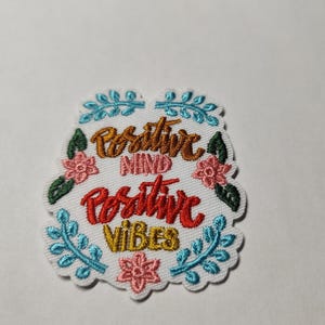 Peut inclure: Un écusson brodé blanc avec le texte Positive Mind en doré, Positive en rouge et Vibes en doré. Le motif est décoré de branches feuillues bleu clair, de fleurs roses et de feuilles vert foncé, le tout entouré d'une bordure festonnée.