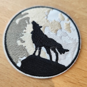 Peut inclure: Écusson brodé circulaire représentant un loup noir hurlant à la lune. La lune est représentée dans des tons de gris et de beige, avec une bordure blanche. Le loup se dresse sur une colline noire, créant un contraste saisissant.