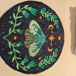 Peut inclure: Patch brodé avec un papillon vert sur fond noir. Le papillon a des ailes orange et vertes et est entouré de feuilles vertes et d'étoiles.