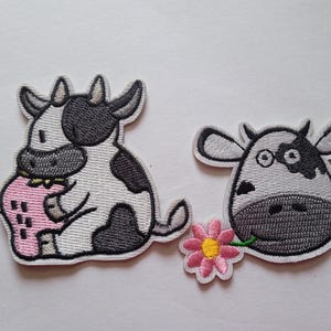 Peut inclure: Deux écussons brodés représentant des vaches de dessin animé. L'une est noire et blanche, tenant une fraise rose. L'autre est une tête de vache avec une fleur dans la bouche. Les écussons ont une bordure blanche.