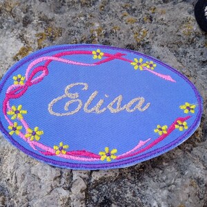 Peut inclure: Écusson brodé ovale avec le nom "Elisa" en fil beige sur fond bleu clair. Le patch est orné d'un ruban rose et de fleurs jaunes, avec une bordure violette.