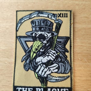 Peut inclure: Écusson brodé représentant un médecin de la peste squelettique avec une faux et un chapeau haut de forme. Le design comprend un triangle et le chiffre romain XIII. Le texte "THE PLAGUE" est en bas, sur fond beige.