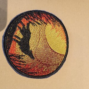 Peut inclure: Patch brodé avec une silhouette noire d'un oiseau volant au-dessus d'un coucher de soleil aux couleurs orange, rouge et jaune.