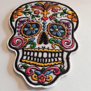 Peut inclure: Patch brodé de sucre blanc avec des détails floraux colorés, y compris des fleurs roses, bleues, vertes et jaunes. Le crâne a des yeux noirs et un nez noir.