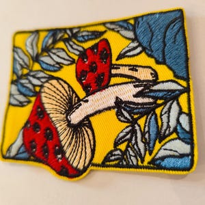 Peut inclure: Écusson brodé avec un motif de champignon. Le patch a un fond jaune vif avec un chapeau de champignon rouge parsemé de points noirs. Le champignon est entouré de feuilles bleues et blanches, le tout souligné en noir.