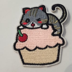 Peut inclure: Écusson brodé représentant un chat tigré gris sur un cupcake rose avec une cerise rouge. Le patch a une bordure noire et est conçu pour être appliqué sur des vêtements ou des accessoires.