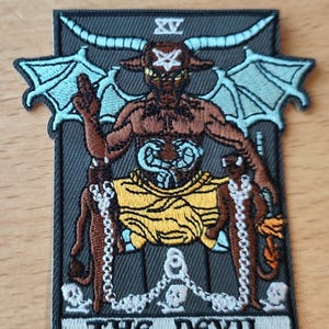 Peut inclure: Écusson brodé représentant Le Diable d'une carte de tarot. L'image montre une figure cornue avec des ailes, assise sur un trône, avec le texte "THE DEVIL" en bas. L'écusson a un fond gris avec des détails marron, jaune et bleu.