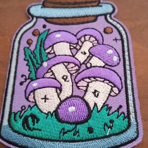 Peut inclure: Écusson brodé représentant un bocal rempli de champignons violets. Le bocal a un fond violet clair avec une bordure turquoise et un couvercle en liège. Les champignons ont des taches blanches et sont posés sur de l'herbe verte.